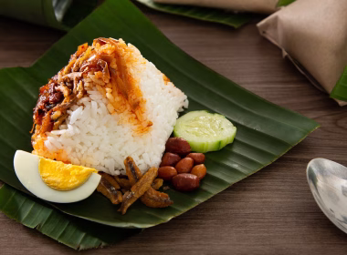 Nasi lemak berbungkus daun pisang dengan sambal, telur rebus, kacang, ikan bilis dan timun dihidang di atas meja kayu.