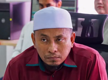 Seorang lelaki berbaju merah dan bersongkok putih sedang duduk dalam suasana pejabat.