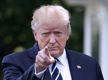 Donald Trump menunjuk ke arah kamera dengan ekspresi serius, mengenakan sut gelap dan tali leher belang.