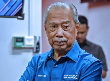 Tan Sri Muhyiddin Yassin memakai baju biru Perikatan Nasional sedang berdiri dengan tangan berpeluk dalam suasana tertutup.