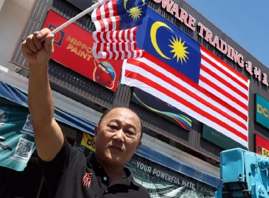Seorang lelaki berdiri di hadapan kedai perkakasan sambil memegang dua bendera kecil Malaysia, dengan Jalur Gemilang gergasi tergantung di bangunan di belakangnya.