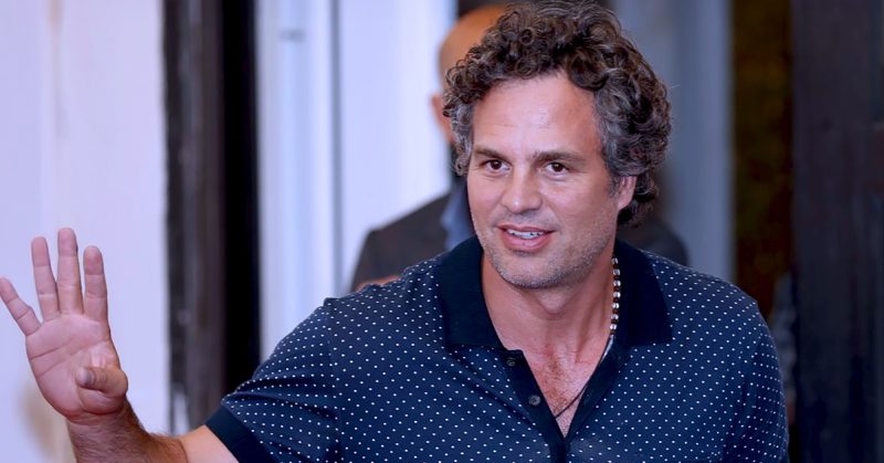 Mark Ruffalo melambai tangan dengan senyuman, memakai kemeja polo biru bercorak titik putih.