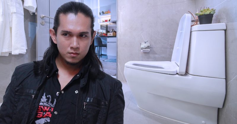 (A) Alt Text: Seorang lelaki berambut panjang berjaket hitam duduk di dalam bilik air moden dengan tandas putih di sebelahnya.