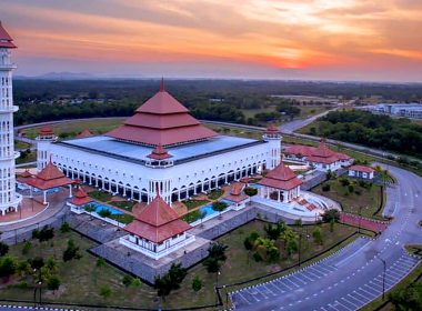 Pemandangan udara Masjid Taman Ilmu di Tembila, Besut, Terengganu dengan menara tinggi dan bumbung merah berlatar matahari terbenam.