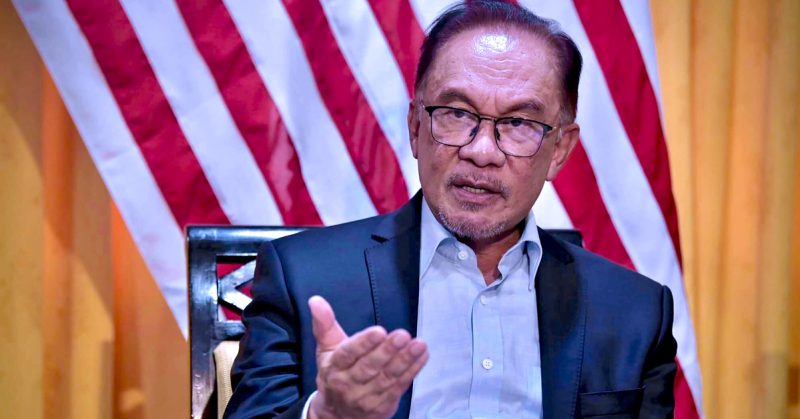 Perdana Menteri Anwar Ibrahim bercakap sambil mengangkat tangan, berlatarbelakangkan Jalur Gemilang.