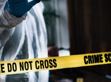 Anggota forensik memakai sut perlindungan lengkap sedang berdiri di belakang pita kuning bertulis “CRIME SCENE DO NOT CROSS” di lokasi kejadian.