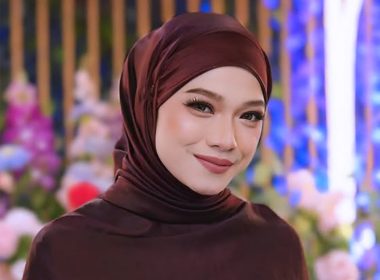 Seorang wanita bertudung maroon tersenyum dengan latar hiasan bunga berwarna-warni.
