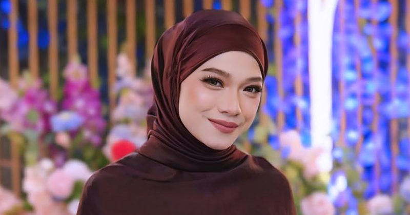 Seorang wanita bertudung maroon tersenyum dengan latar hiasan bunga berwarna-warni.