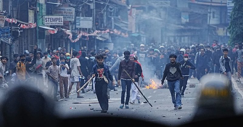 Sekumpulan mahasiswa Indonesia berarak di jalan raya sambil membawa kayu, asap gas pemedih mata dan api kelihatan di latar belakang.