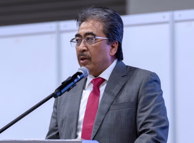 Johari Abdul Ghani berucap di podium dengan memakai sut kelabu dan tali leher merah.