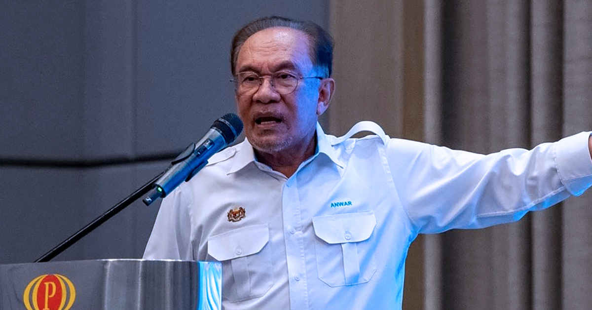 Perdana Menteri Malaysia berucap di podium sambil mengangkat tangan, menjelaskan isu lawatan luar negara atas tuntutan kerja.