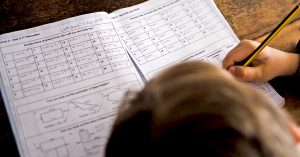 Budak lelaki sedang menulis kerja rumah matematik dalam buku latihan, memegang pensil di atas meja kayu.