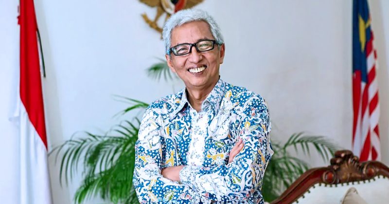Duta Besar Indonesia ke Malaysia tersenyum sambil berpeluk tubuh, berlatarbelakangkan bendera Indonesia dan Malaysia.