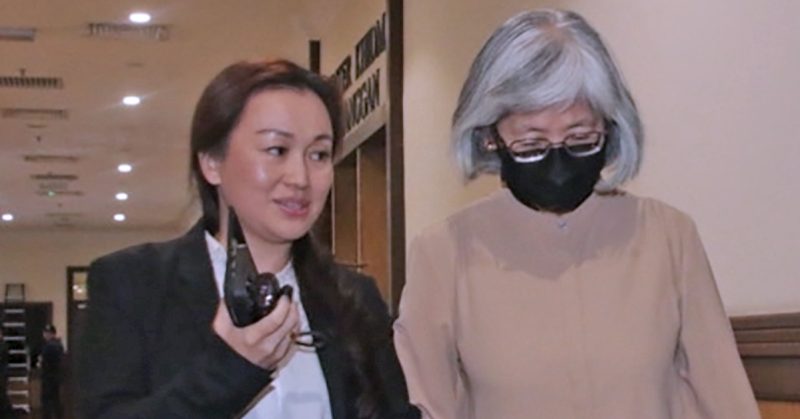 Dua wanita berjalan di koridor mahkamah; seorang berambut hitam memegang walkie-talkie, seorang lagi berambut kelabu memakai pelitup muka hitam.