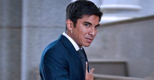 Syed Saddiq dalam sut biru gelap sedang melangkah, wajah bersemangat dengan latar belakang bangunan mahkamah.