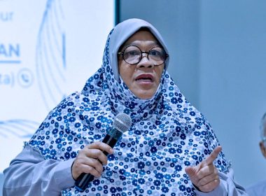 Seorang wanita berhijab bercorak bunga sedang memegang mikrofon dan bercakap ketika sidang media mengenai projek pembangunan semula Kampung Sungai Baru.