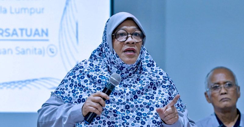 Seorang wanita berhijab bercorak bunga sedang memegang mikrofon dan bercakap ketika sidang media mengenai projek pembangunan semula Kampung Sungai Baru.