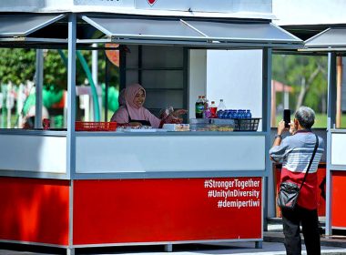 Seorang peniaga wanita berada di dalam kios MyKiosk sambil tersenyum, manakala seorang lelaki mengambil gambar menggunakan telefon bimbit.