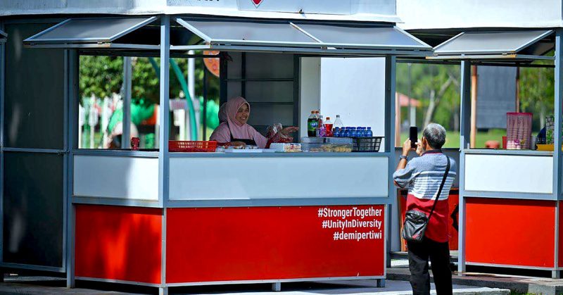 Seorang peniaga wanita berada di dalam kios MyKiosk sambil tersenyum, manakala seorang lelaki mengambil gambar menggunakan telefon bimbit.
