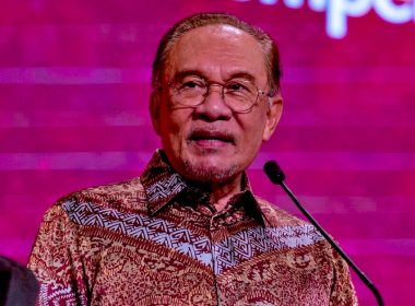 Perdana Menteri Anwar Ibrahim berucap di majlis rasmi dengan latar merah, memakai baju batik.