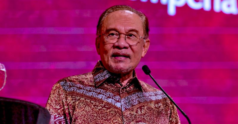 Perdana Menteri Anwar Ibrahim berucap di majlis rasmi dengan latar merah, memakai baju batik.