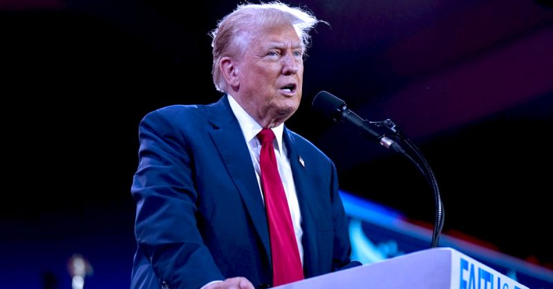 Donald Trump berucap di podium dengan mikrofon, mengenakan sut biru gelap dan tali leher merah.