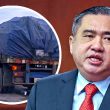 Anthony Loke, Menteri Pengangkutan Malaysia, bercakap di hadapan; di sebelahnya imej lori sarat muatan ditutup kanvas.