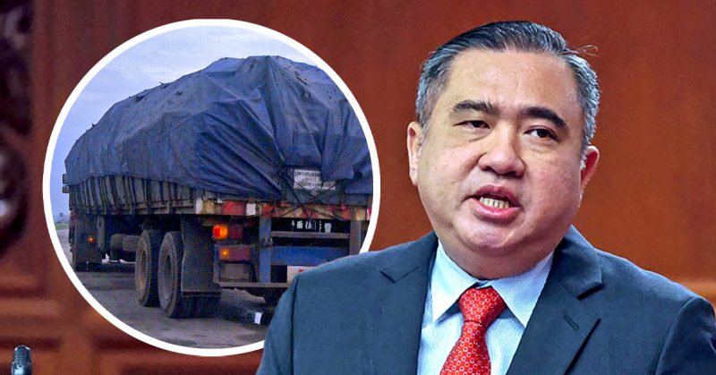 Anthony Loke, Menteri Pengangkutan Malaysia, bercakap di hadapan; di sebelahnya imej lori sarat muatan ditutup kanvas.