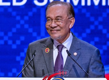 Perdana Menteri Anwar Ibrahim berdiri di podium dengan dua mikrofon, tersenyum di hadapan latar biru.
