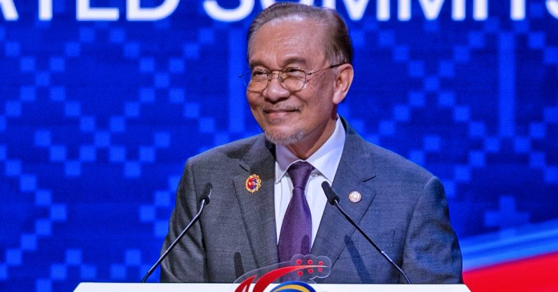 Perdana Menteri Anwar Ibrahim berdiri di podium dengan dua mikrofon, tersenyum di hadapan latar biru.