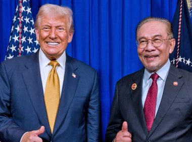 Donald Trump dan Anwar Ibrahim berpakaian sut formal berdiri bersebelahan di hadapan latar biru dengan bendera di belakang.