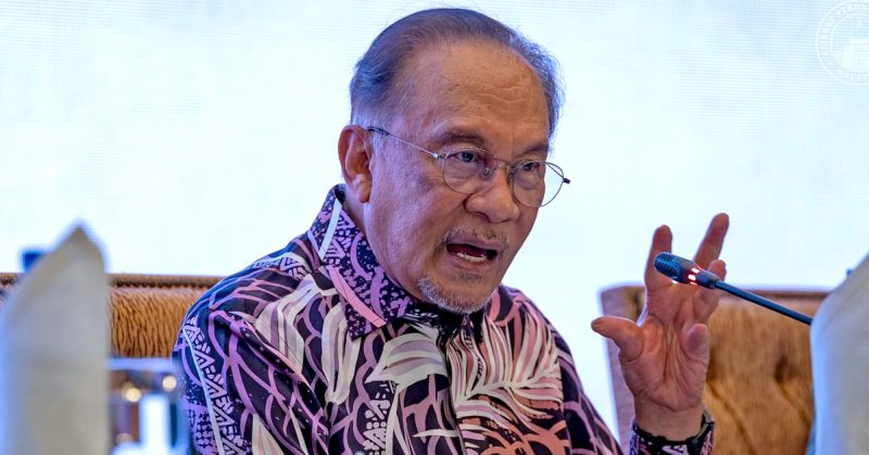 Perdana Menteri Malaysia, Anwar Ibrahim sedang bercakap di forum dengan ekspresi tegas, memakai baju batik bercorak ungu dan hitam.