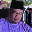 Dr MAZA dalam pakaian ungu sedang memberi isyarat tangan, dengan imej sisipan Donald Trump disambut meriah oleh pegawai Malaysia di lapangan terbang.