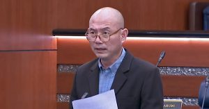 Ahli Parlimen berdiri di Dewan Rakyat sambil memegang kertas ucapan semasa sesi perbahasan, latar dewan parlimen Malaysia.