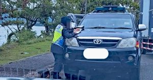 Pegawai JPJ wanita sedang menulis saman pada sebuah kenderaan Toyota hitam di tepi jalan, berkaitan kes pemandu e-hailing disaman kerana baju tidak berkolar.
