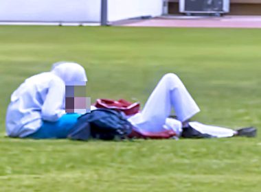 Dua individu berpakaian sekolah berada di atas padang awam; seorang duduk menunduk, seorang lagi berbaring sambil beg sekolah diletakkan di sisi.