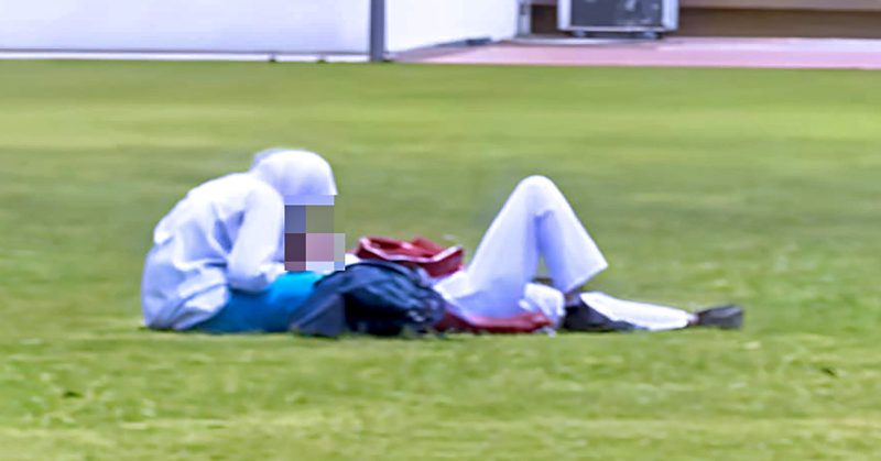 Dua individu berpakaian sekolah berada di atas padang awam; seorang duduk menunduk, seorang lagi berbaring sambil beg sekolah diletakkan di sisi.