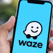 Tangan memegang telefon pintar yang memaparkan logo aplikasi navigasi Waze pada skrin dengan latar belakang jalan raya.