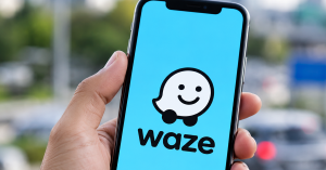 Tangan memegang telefon pintar yang memaparkan logo aplikasi navigasi Waze pada skrin dengan latar belakang jalan raya.